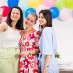 Three women joyfully taking a selfie against a colorful balloon backdrop indoors.